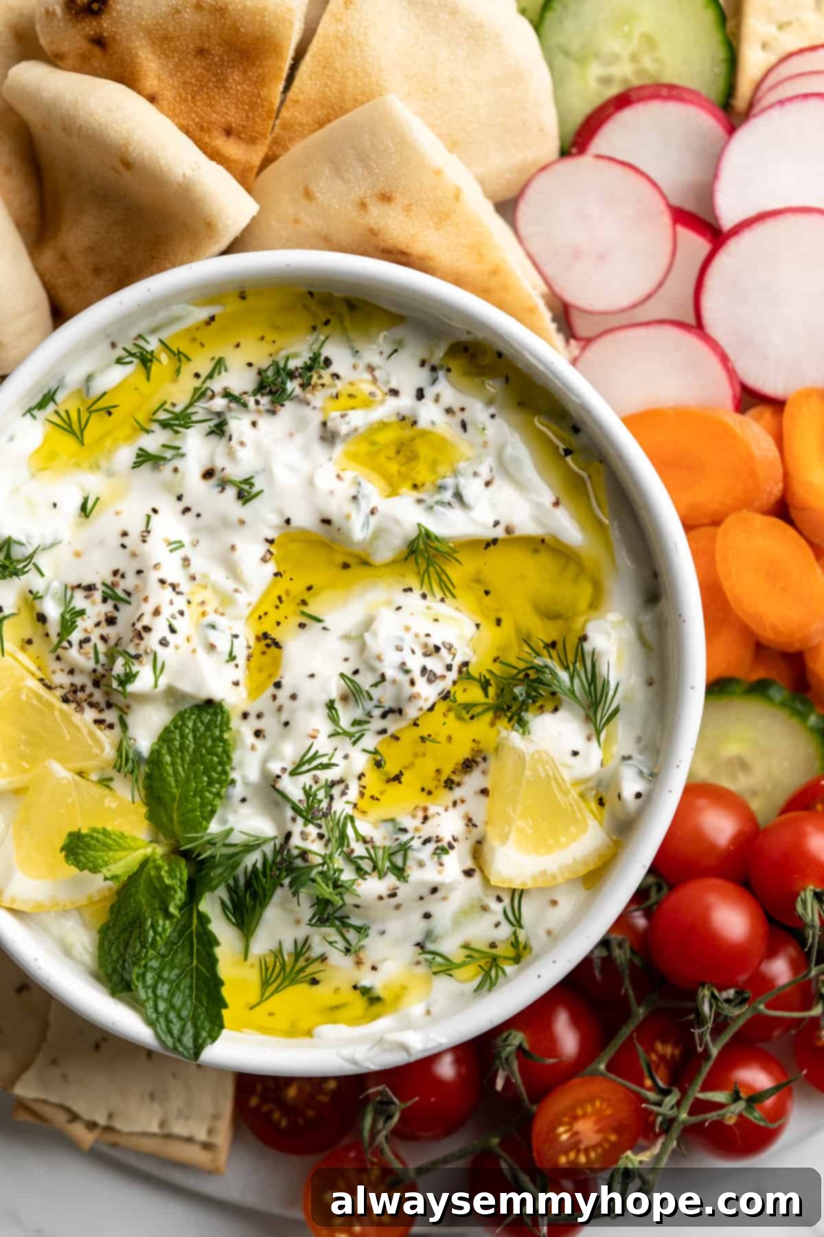 Homemade vegan tzatziki is a tangy, refreshing, and garlicky dip with bright lemon flavor. This easy plant-based recipe is perfect for any meal! An overhead view of a bowl of freshly made vegan tzatziki, garnished with a drizzle of olive oil, fresh herbs, and lemon slices.