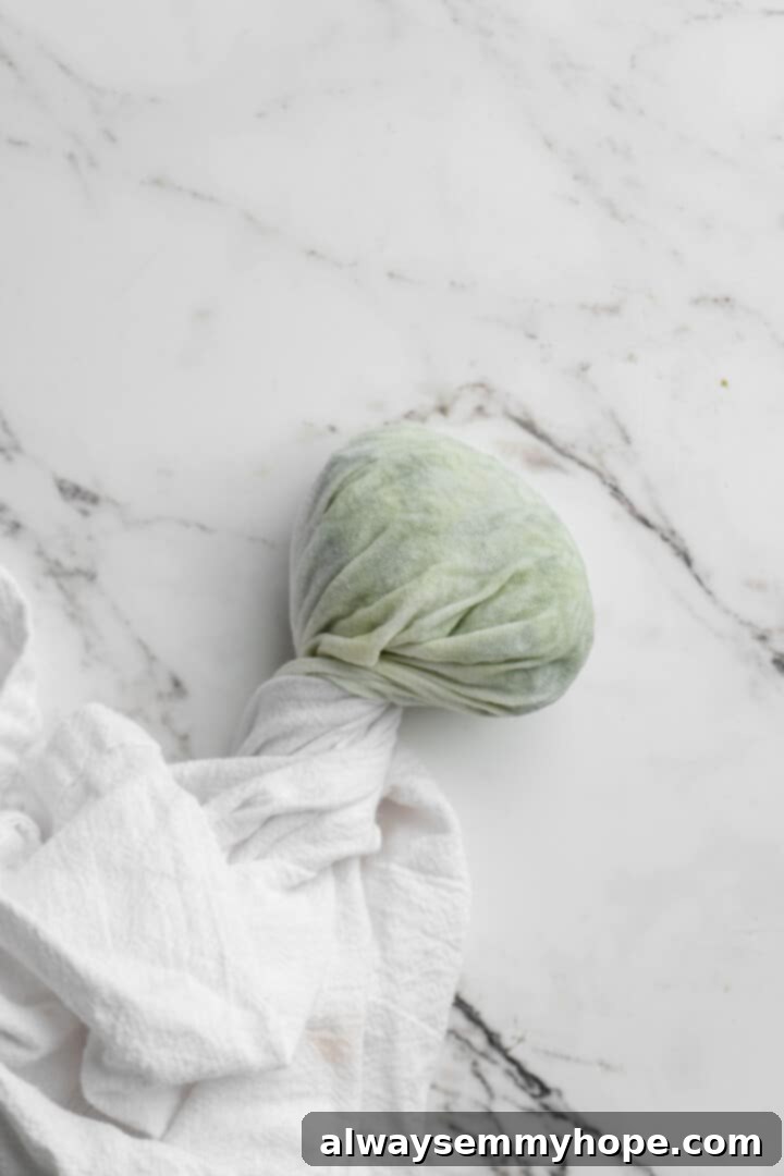 Homemade vegan tzatziki is a tangy, refreshing, and garlicky dip with bright lemon flavor. This easy plant-based recipe is perfect for any meal! An overhead shot of grated cucumber tightly wrapped in a white kitchen towel, with moisture visibly being squeezed out over a sink.