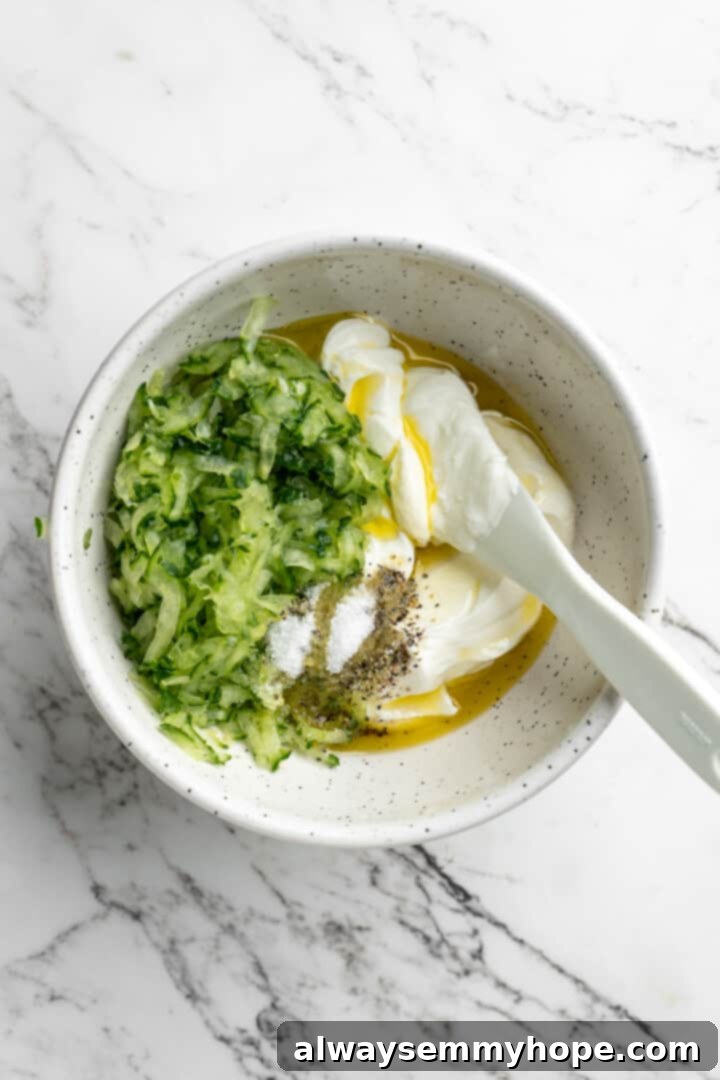 Homemade vegan tzatziki is a tangy, refreshing, and garlicky dip with bright lemon flavor. This easy plant-based recipe is perfect for any meal! An overhead shot of a mixing bowl containing all the ingredients for vegan tzatziki – squeezed cucumber, vegan Greek yogurt, garlic, lemon juice, olive oil, salt, and pepper – with a spoon poised for stirring.