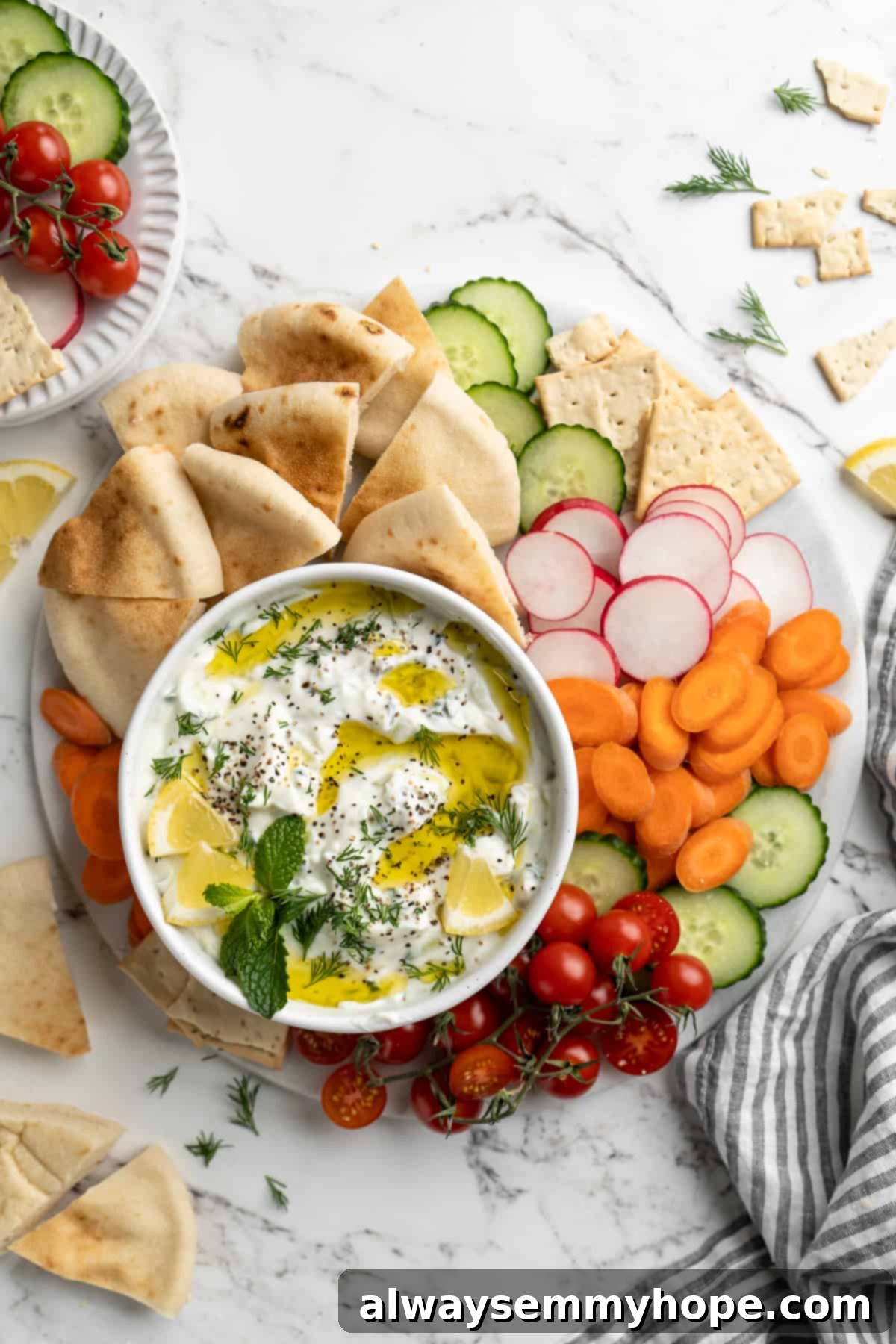 Homemade vegan tzatziki is a tangy, refreshing, and garlicky dip with bright lemon flavour. This easy plant-based recipe is perfect for any meal! An overhead view of a bowl of vegan tzatziki presented on a large platter, surrounded by an appetizing spread of fresh vegetables, warm pita bread, and crispy crackers.