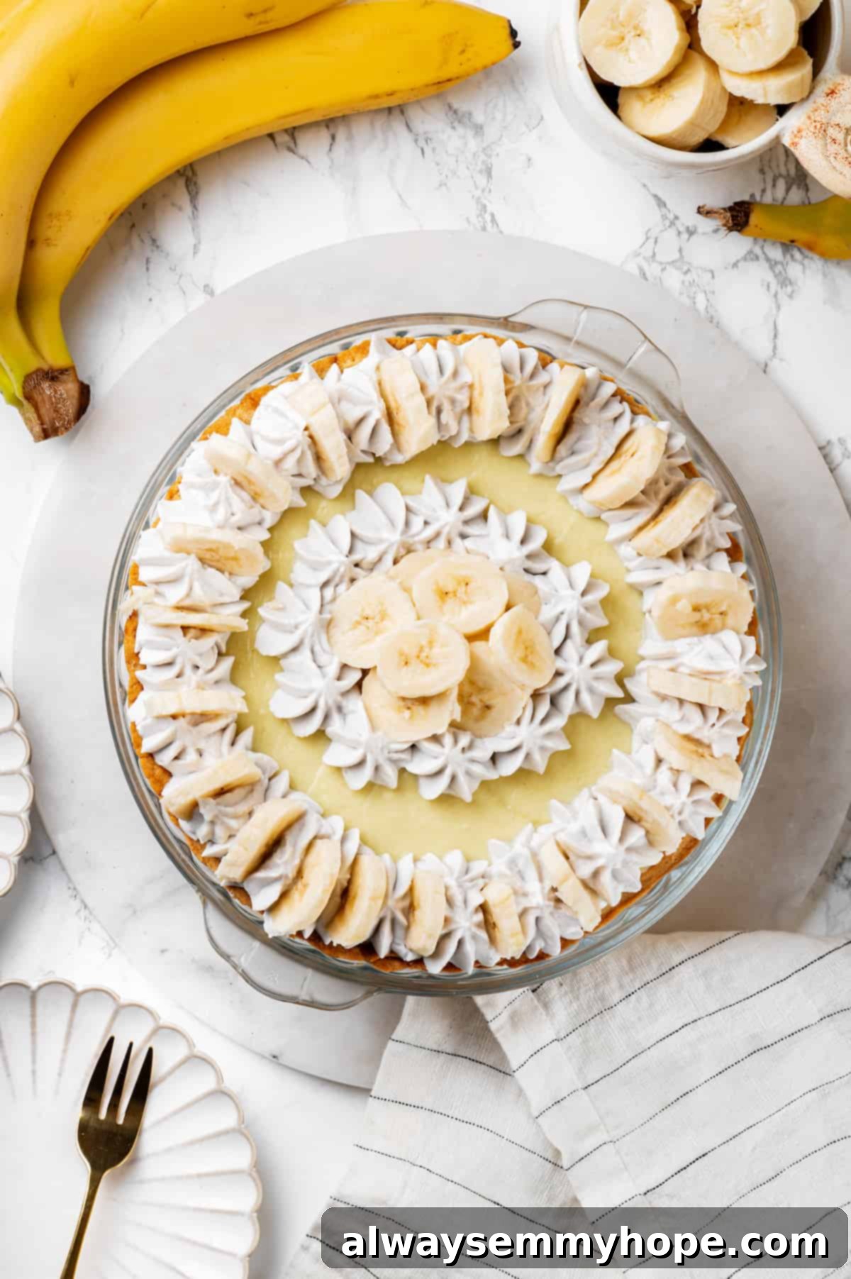 Overhead view of a stunning whole vegan banana cream pie, freshly topped with coconut whipped cream and banana slices.