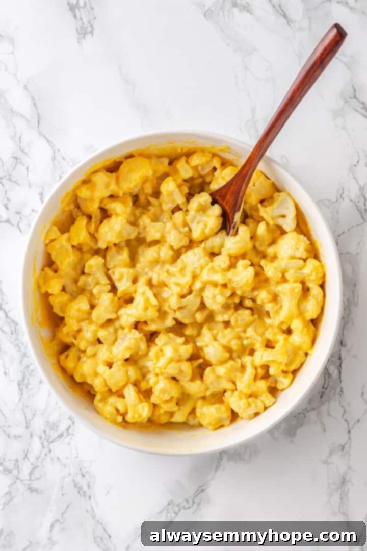 Cooked cauliflower coated in vegan cheese sauce, stirred in a large mixing bowl with a wooden spoon.