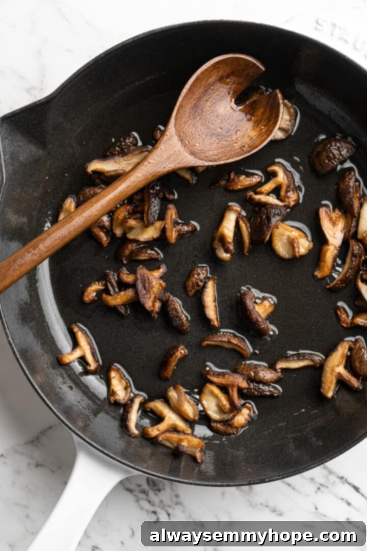 Overhead view of shiitakes cooking in skillet with wooden spoon, becoming golden brown and tender.