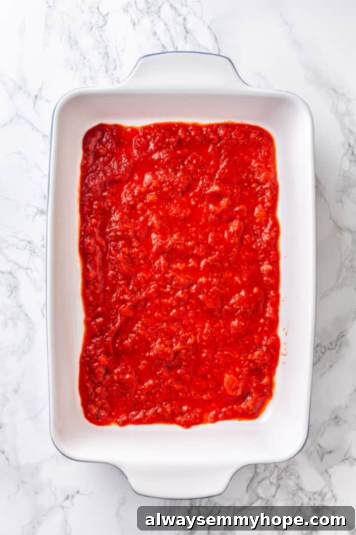 Overhead view of a thin, even layer of marinara sauce spread across the bottom of a blue ceramic baking dish.