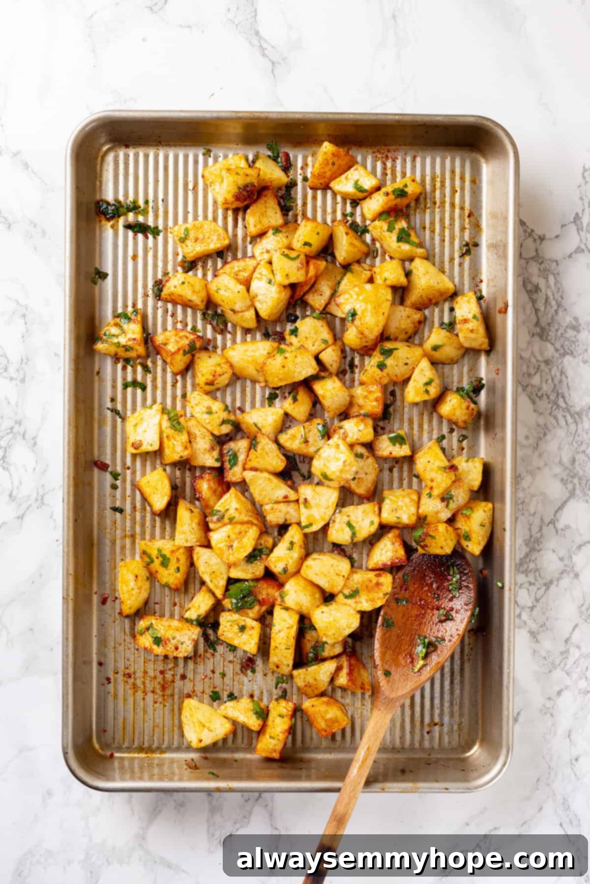 Overhead view of Batata Harra on a baking sheet, freshly tossed with the green spicy sauce and a wooden spoon nearby.