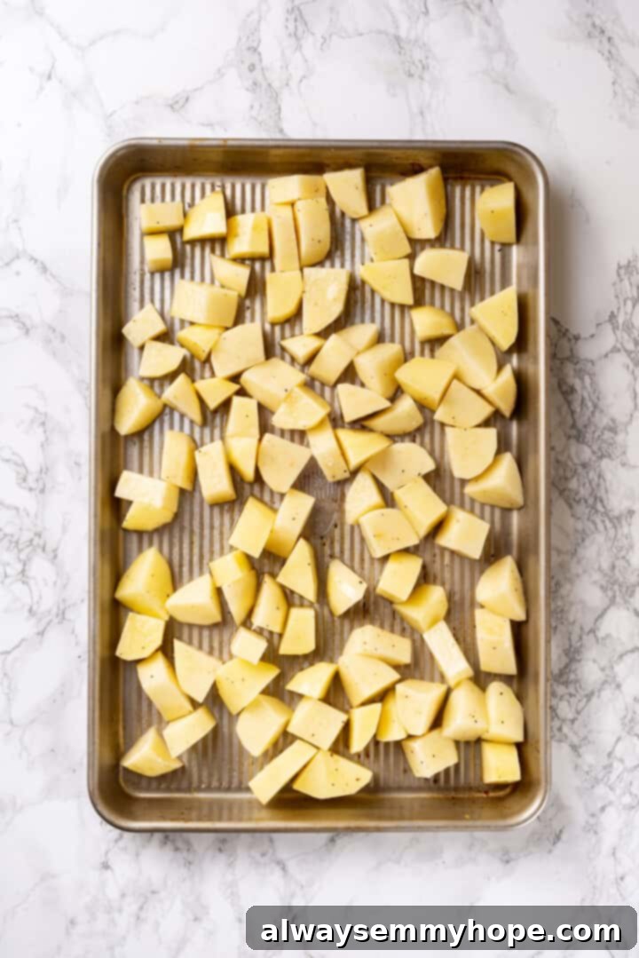 Overhead view of seasoned potato cubes spread evenly on a baking sheet, ready for oven roasting.