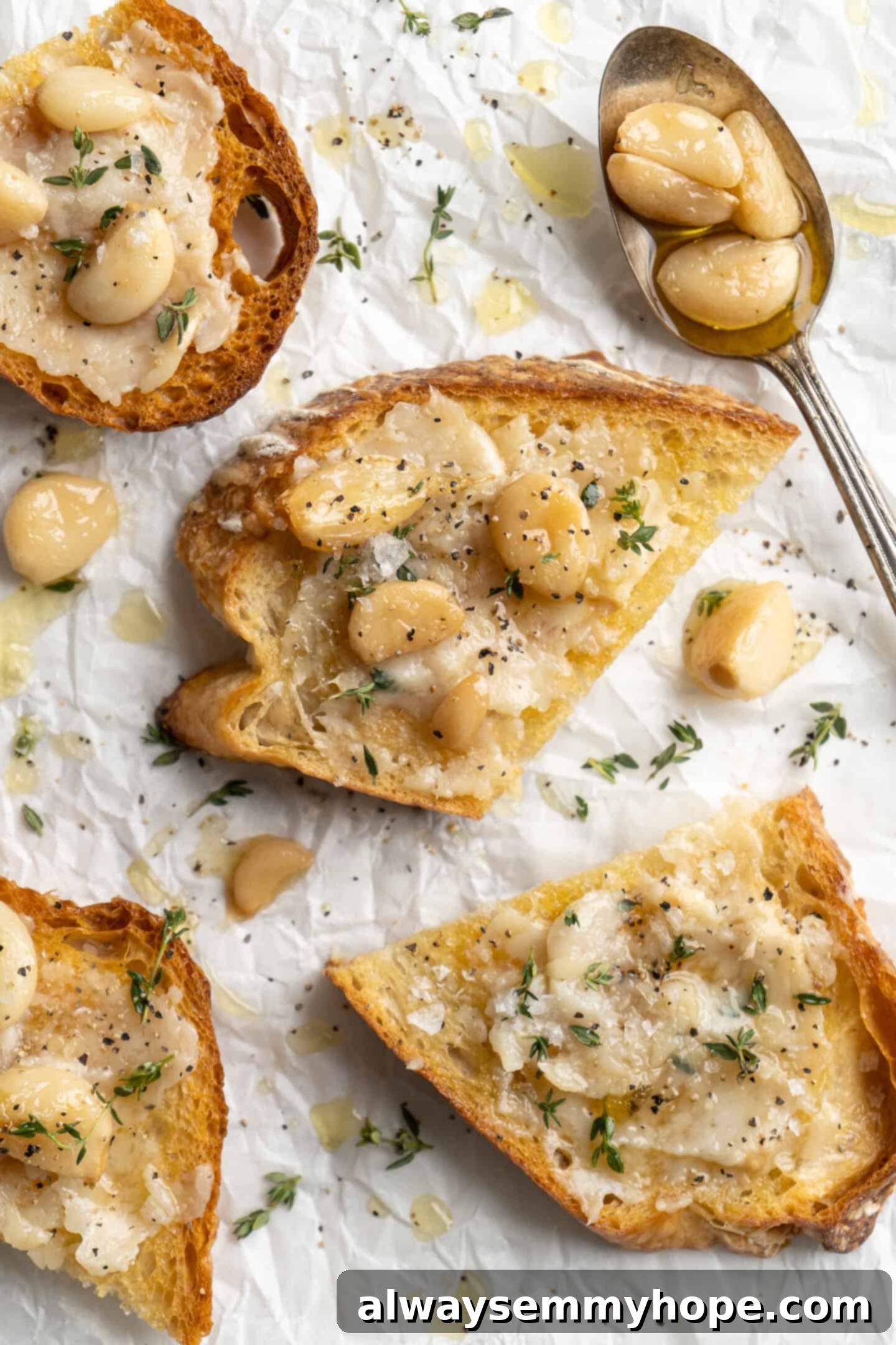 Overhead view of garlic confit on crusty toast with fresh thyme, black pepper, and flaky sea salt. The golden-brown garlic cloves are mashed slightly into the toast, sprinkled with herbs and seasoning.