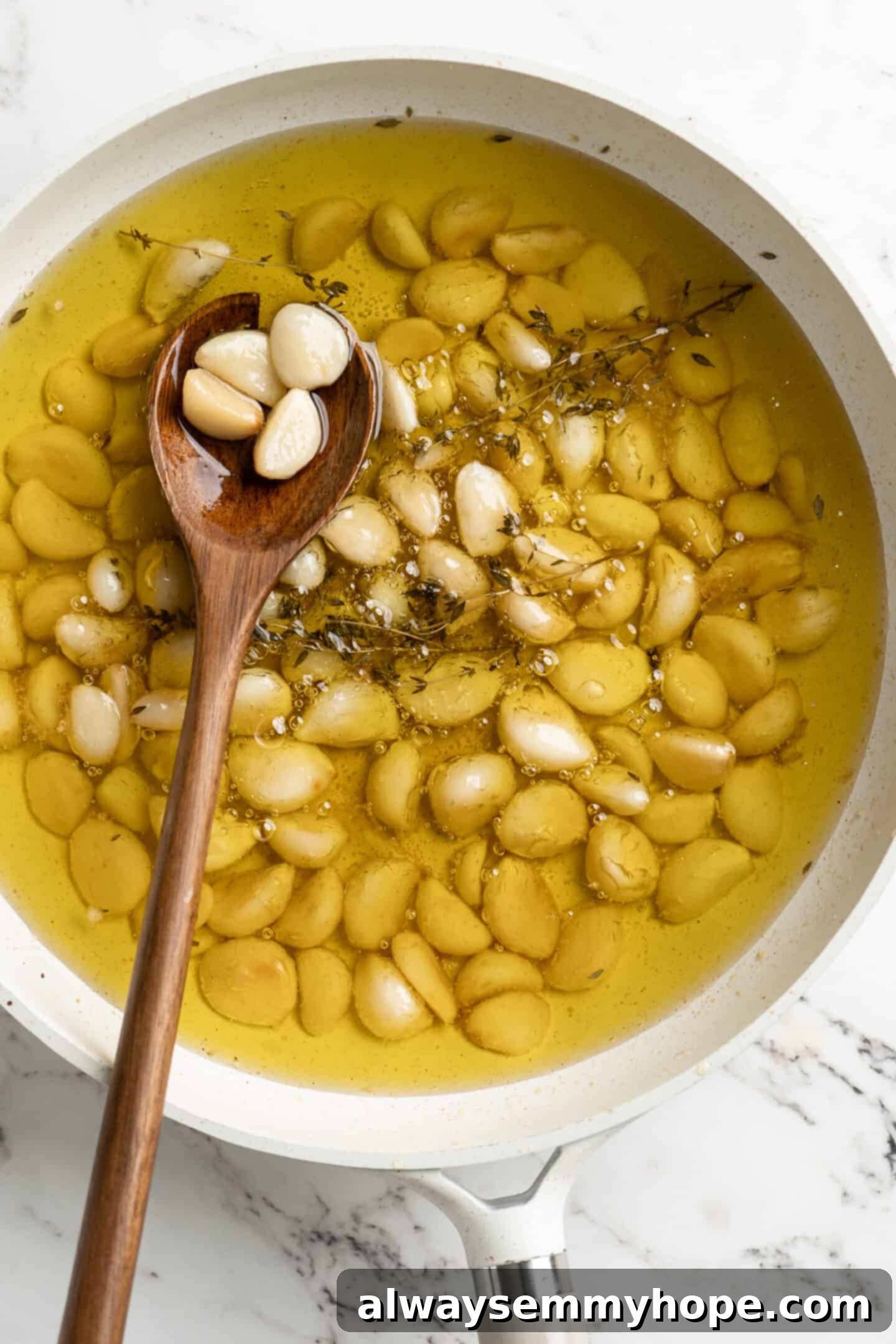 Overhead view of garlic confit in a pan after cooking, with a wooden spoon resting inside, showcasing the soft, golden cloves.