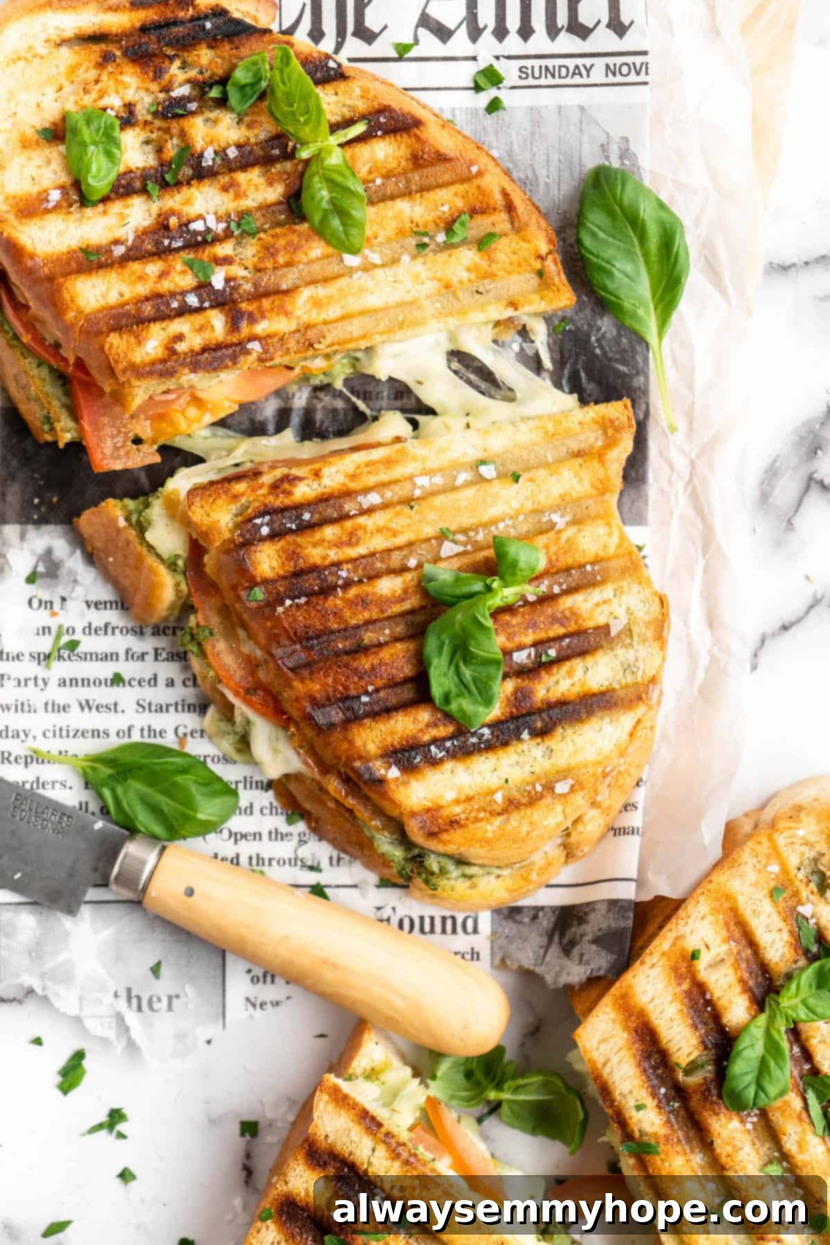 Halved vegan caprese sandwiches set on newspaper, with basil leaves scattered over them for garnish