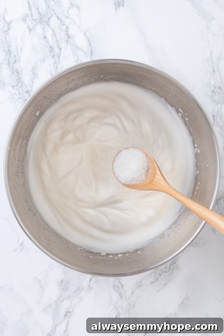 Overhead view of sugar being added to bowl of whipped aquafaba
