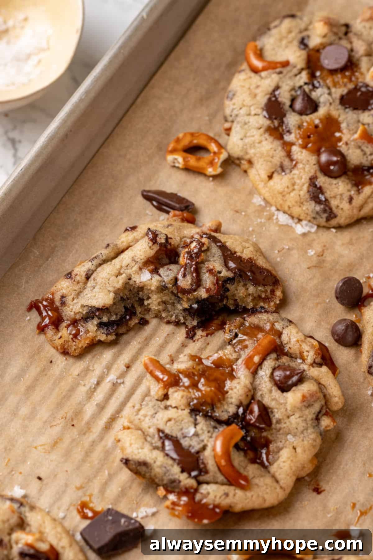 With an irresistible mix of chocolate, caramel and crispy pretzels, these kitchen sink cookies are easy to whip up and even easier to devour! Vegan kitchen sink cookies on a parchment-lined baking sheet, with one cookie broken in half to show its interior.