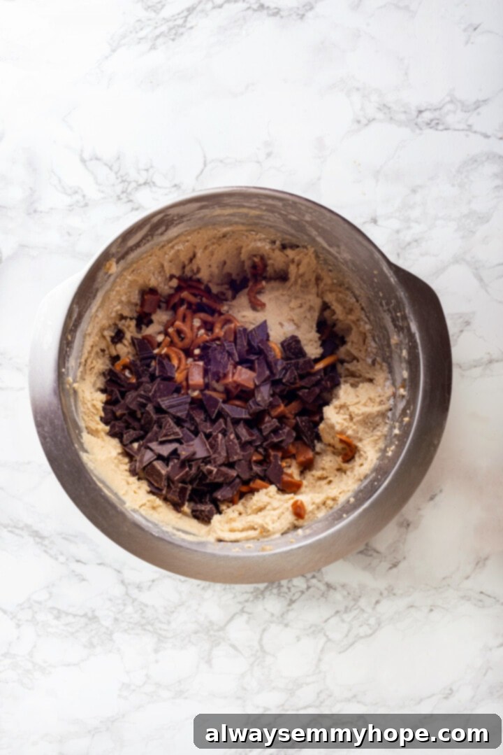 With an irresistible mix of chocolate, caramel and crispy pretzels, these kitchen sink cookies are easy to whip up and even easier to devour! Overhead view of chocolate chunks, chips, pretzels, and chopped caramels being added to the cookie dough in a mixing bowl.