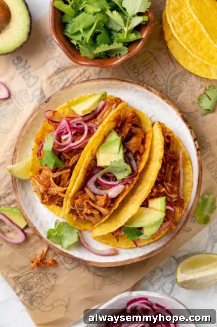 Overhead view of 3 jackfruit tacos on plate surrounded by tortillas, bowl of cilantro, and other toppings