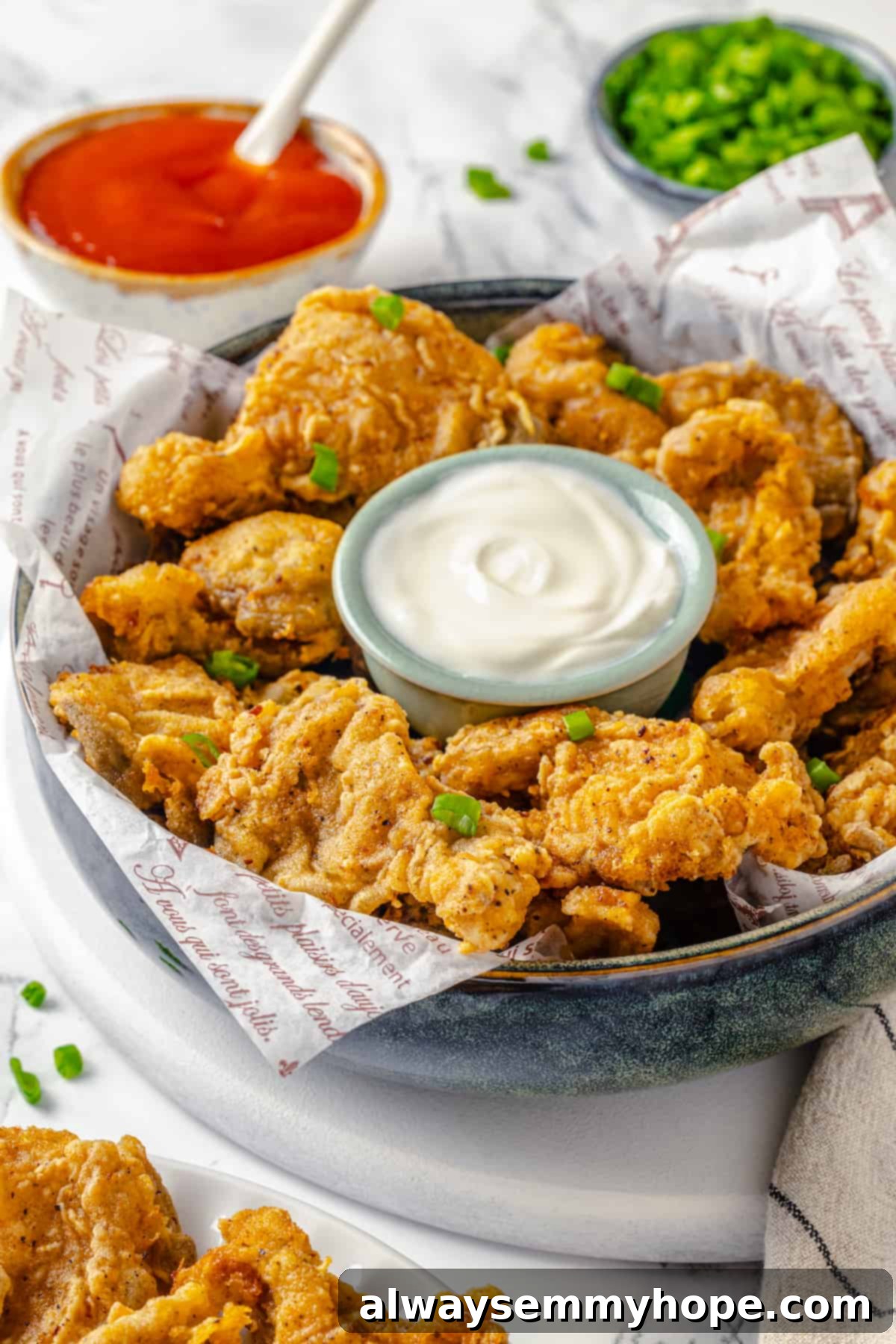 This vegan fried chicken is crispy, delicious, and perfect with a creamy dipping sauce. A bowl of vegan fried chicken with a small dish of creamy dipping sauce in the center and a bottle of hot sauce blurred in the background.