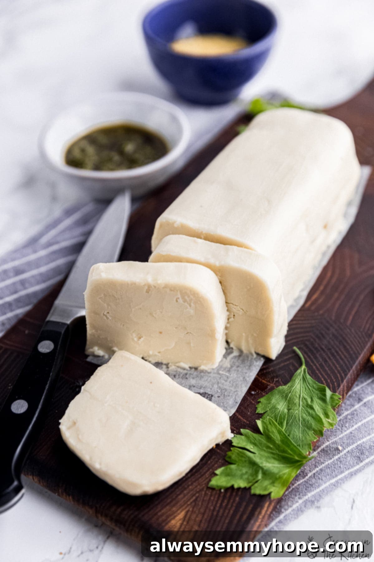 vegan butter dairy substitute in a log with a few pieces sliced, on a brown cutting board surrounded by condiments and parsley and a knife and crackers