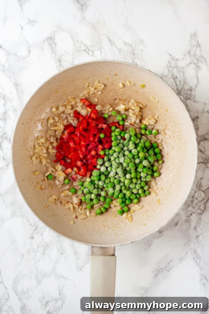 Overhead view of frozen peas and red bell pepper added to pan of cooked onions