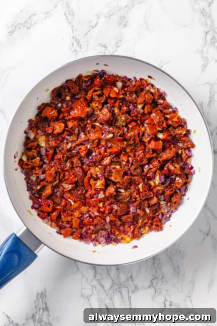 Sautéing sun-dried tomatoes, red onions, and garlic for the creamy pasta sauce base. Sun-dried tomatoes and red onions sautéing in a skillet with sun-dried tomato oil and vegan butter, creating a flavorful base.
