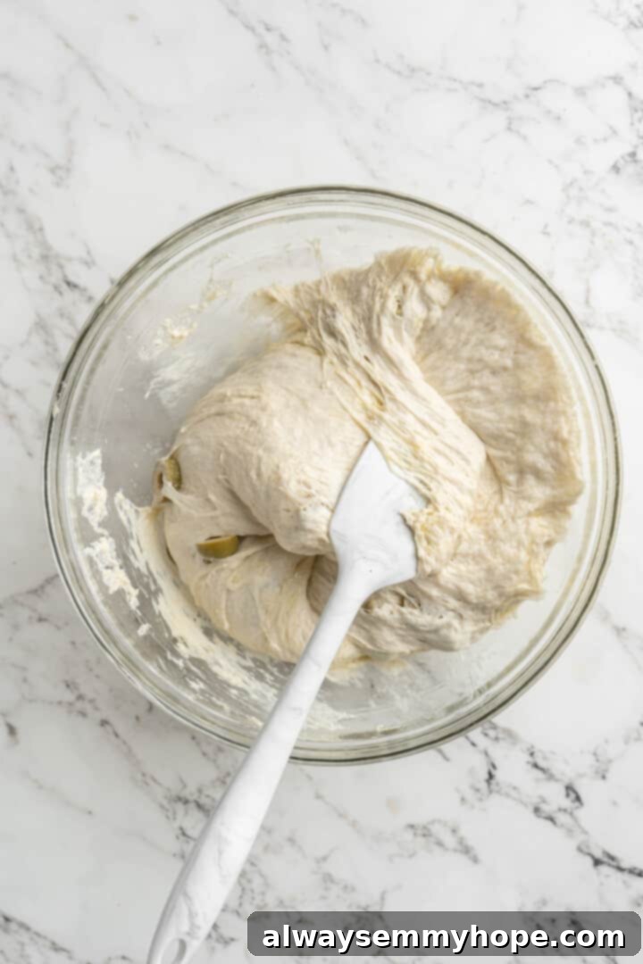 This crusty no-knead olive bread recipe is perfect for beginning bakers, but seasoned pros will love it too. Easy and delicious! Overhead view of spatula continuing to fold dough in a glass bowl, building tension