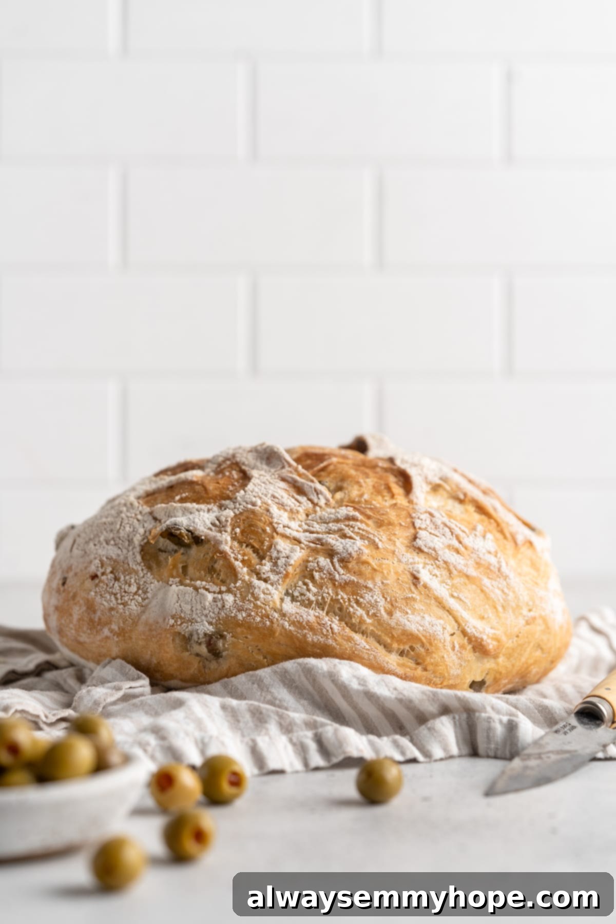 This crusty no-knead olive bread recipe is perfect for beginning bakers, but seasoned pros will love it too. Easy and delicious! Whole olive bread loaf resting on a tea towel, ready for serving