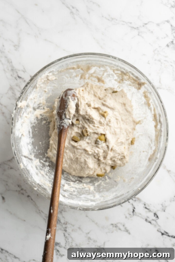 This crusty no-knead olive bread recipe is perfect for beginning bakers, but seasoned pros will love it too. Easy and delicious! Overhead view of wooden spoon continuing to stir dough for olive bread, showing its shaggy texture