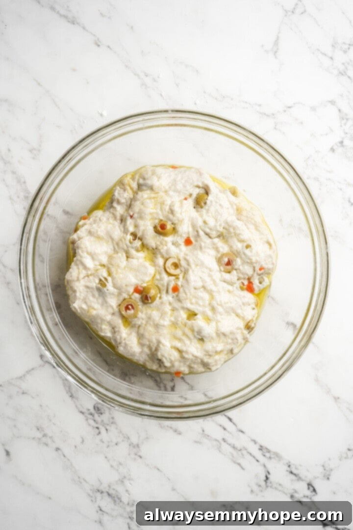 This crusty no-knead olive bread recipe is perfect for beginning bakers, but seasoned pros will love it too. Easy and delicious! Olive bread dough resting in a glass bowl, lightly coated with oil and covered