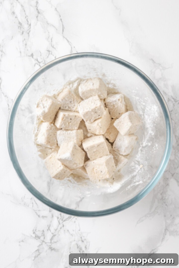 Overhead view of cubed tofu being tossed in a cornstarch, salt, and pepper mixture in a mixing bowl
