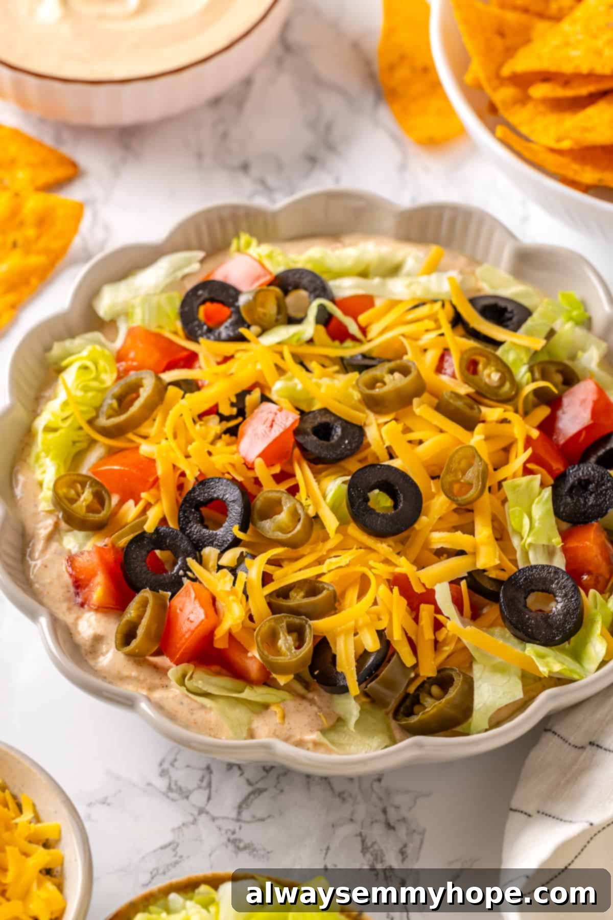 This easy vegan taco dip recipe features a creamy, cheesy taco-seasoned base topped with fresh lettuce, vegan cheese, juicy tomato, and black olives. It's completely plant-based and perfect for any party! Serving bowl with colorful vegan taco dip, garnished with fresh ingredients and ready to eat