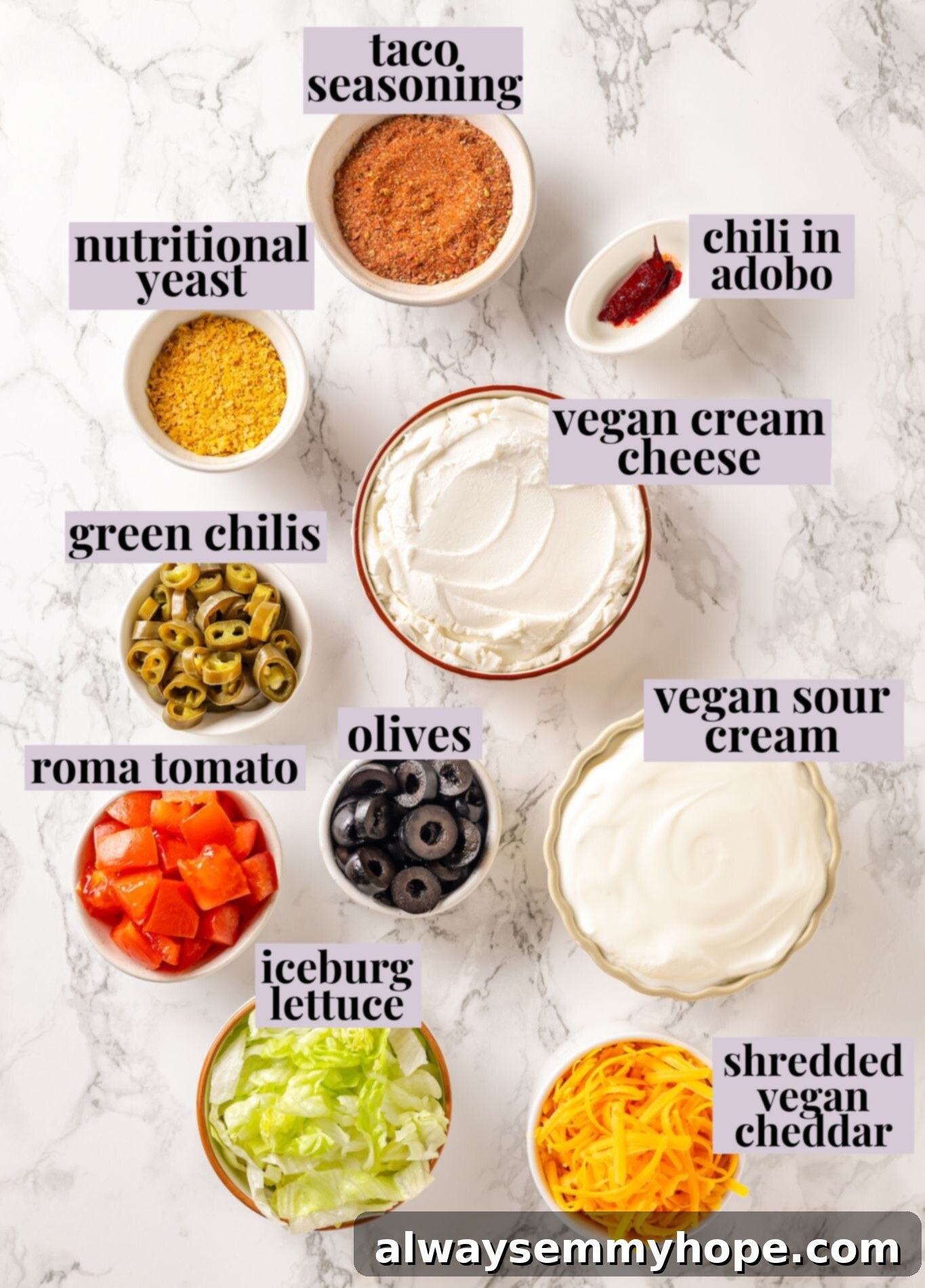 Fresh ingredients laid out for making this easy vegan taco dip recipe, including vegan sour cream, cream cheese, taco seasoning, lettuce, tomatoes, and vegan cheddar cheese. Overhead view of fresh ingredients for making homemade vegan taco dip laid out on a surface