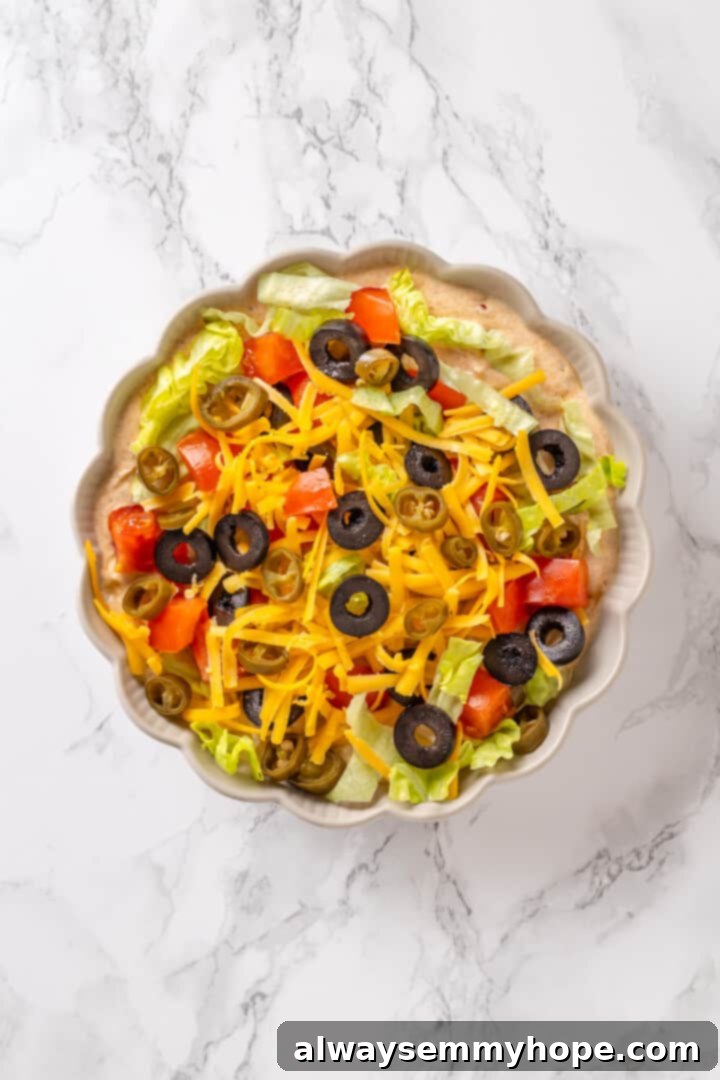 Serve your easy vegan taco dip by topping the creamy base with fresh lettuce, vegan cheddar cheese, tomato, green chilis, and black olives. Assembled vegan taco dip with all colorful toppings arranged in a bowl, ready to serve
