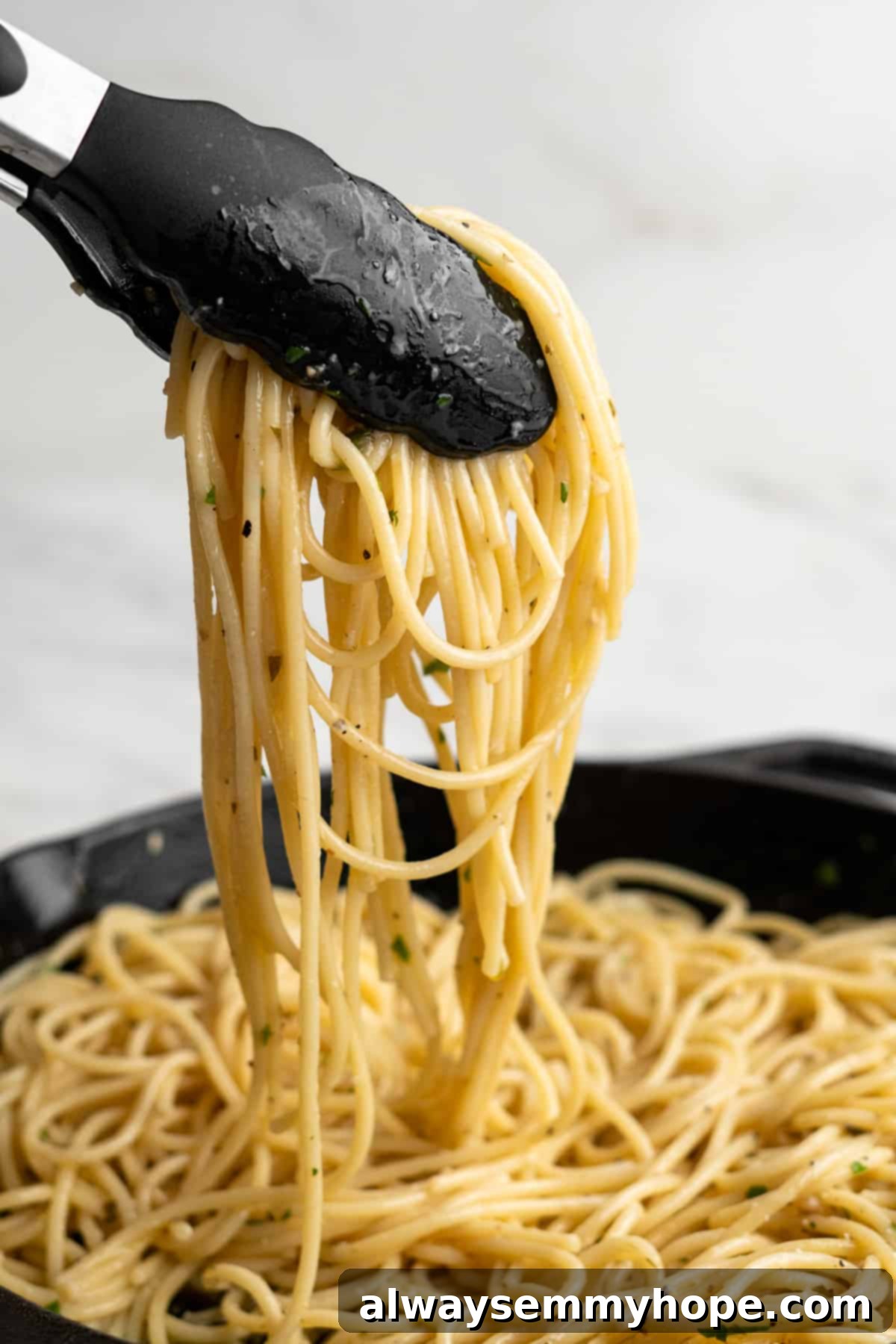This lemon pasta recipe is light and flavourful, with bright lemon, plenty of garlic, and umami vegan Parmesan. An easy weeknight dinner! Tongs pulling a serving of zesty lemon pasta from a skillet, showing the perfectly coated noodles.