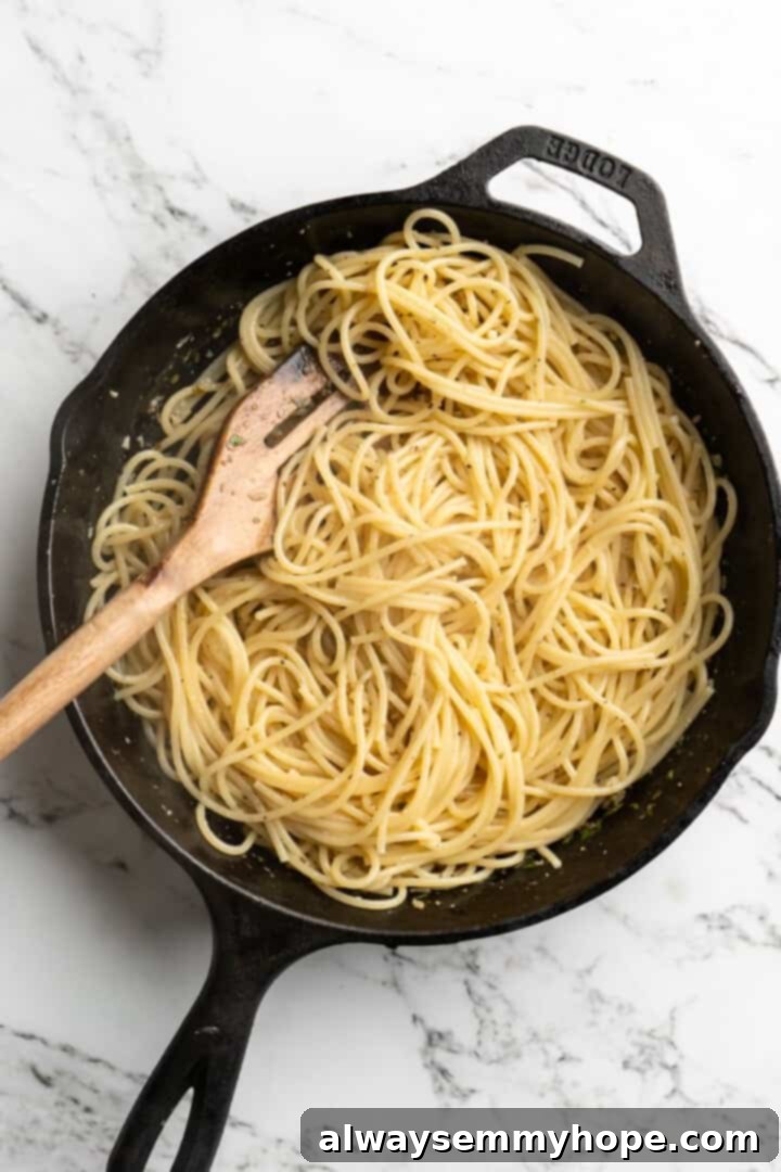 This lemon pasta recipe is light and flavourful, with bright lemon, plenty of garlic, and umami vegan Parmesan. An easy weeknight dinner! Overhead view of a wooden spatula stirring the lemon pasta in a skillet, ensuring the sauce evenly coats the noodles.