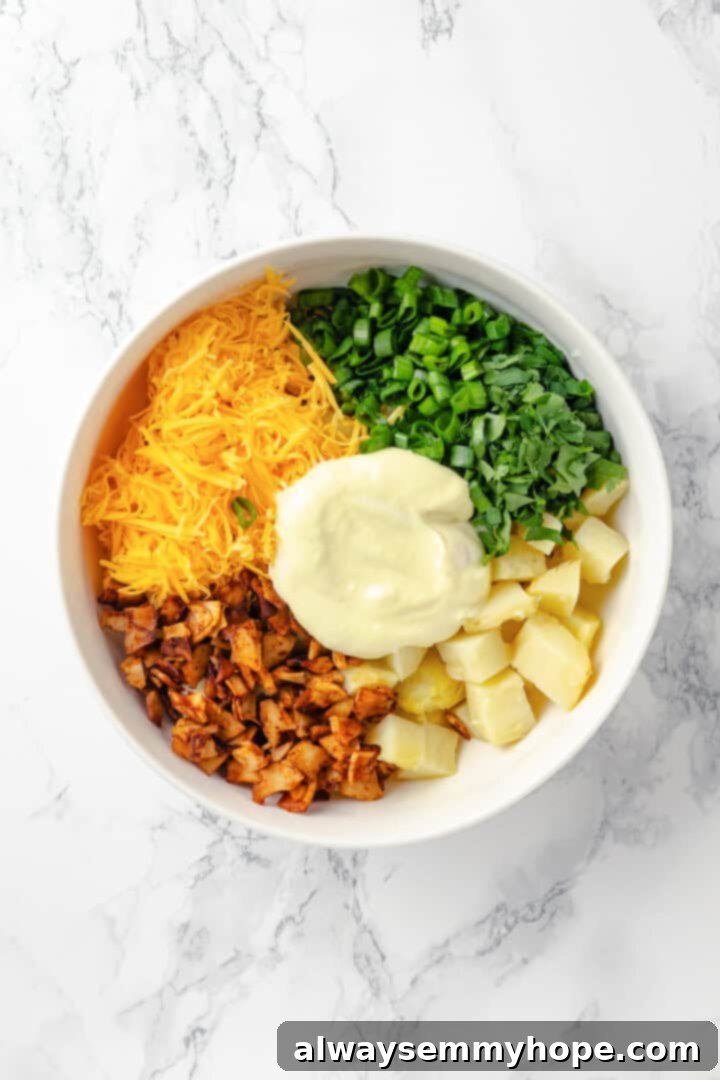 Overhead shot of cubed baked potatoes, chopped scallions, shredded vegan cheddar cheese, and vegan bacon crumbles in a large mixing bowl, awaiting the creamy dressing.