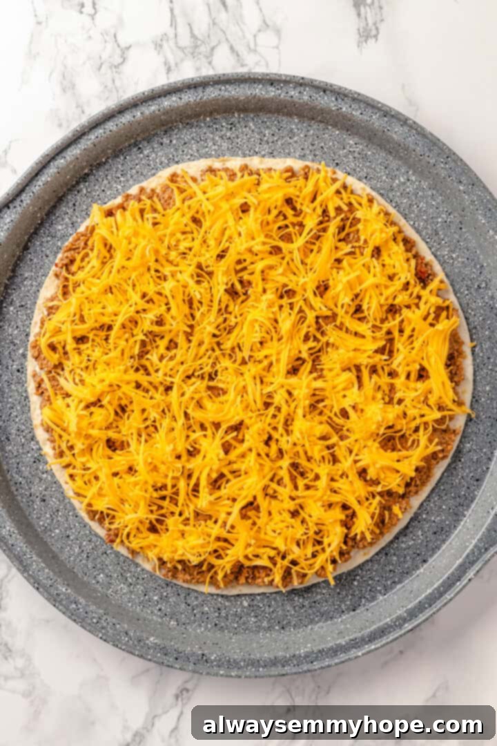 With layers of seasoned taco meat, plant-based cheese, and tortillas, this easy taco bake recipe is sure to please the whole family! Overhead view of the first assembled layer of the taco bake, showing a tortilla topped with seasoned vegan meat and shredded cheese in a round pan.