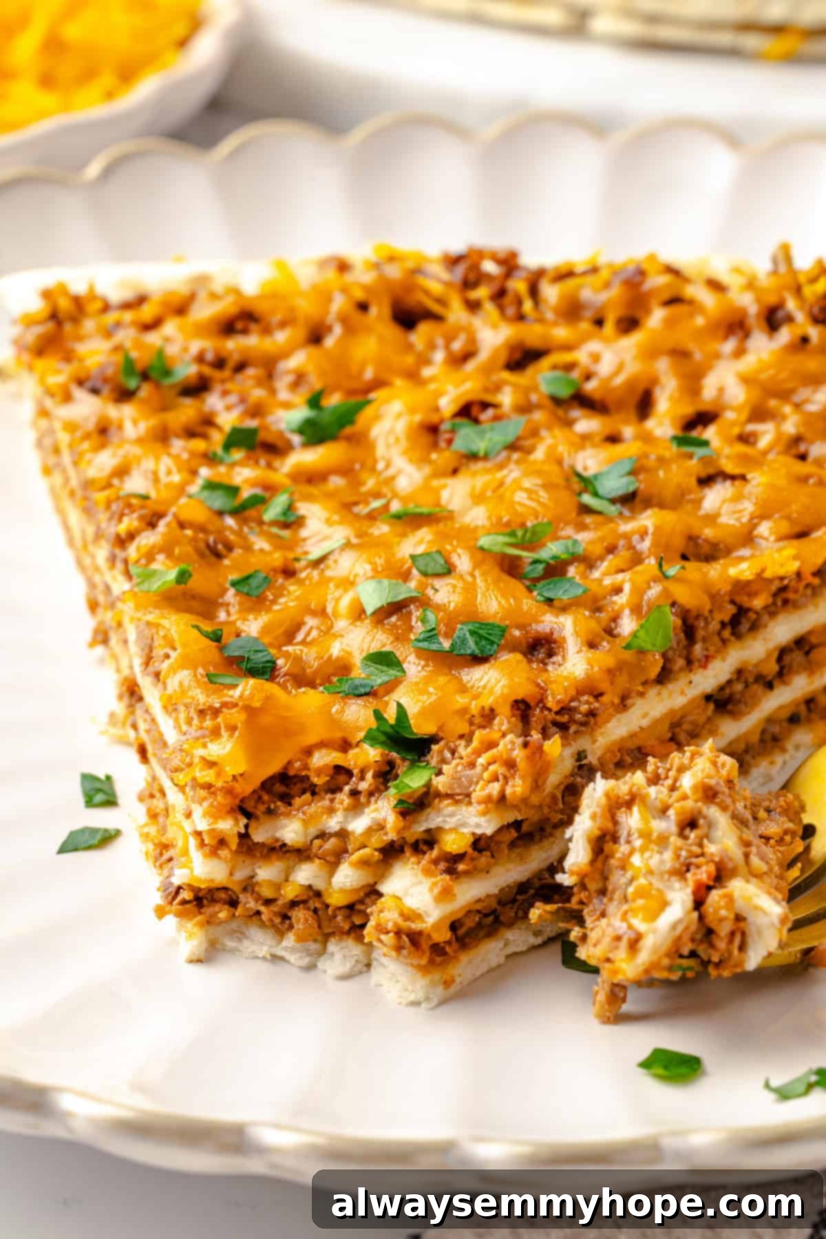 With layers of seasoned taco meat, plant-based cheese, and tortillas, this easy taco bake recipe is sure to please the whole family! Close-up shot of a slice of vegan taco bake on a plate, with a fork digging into the top, showing the texture and layers.