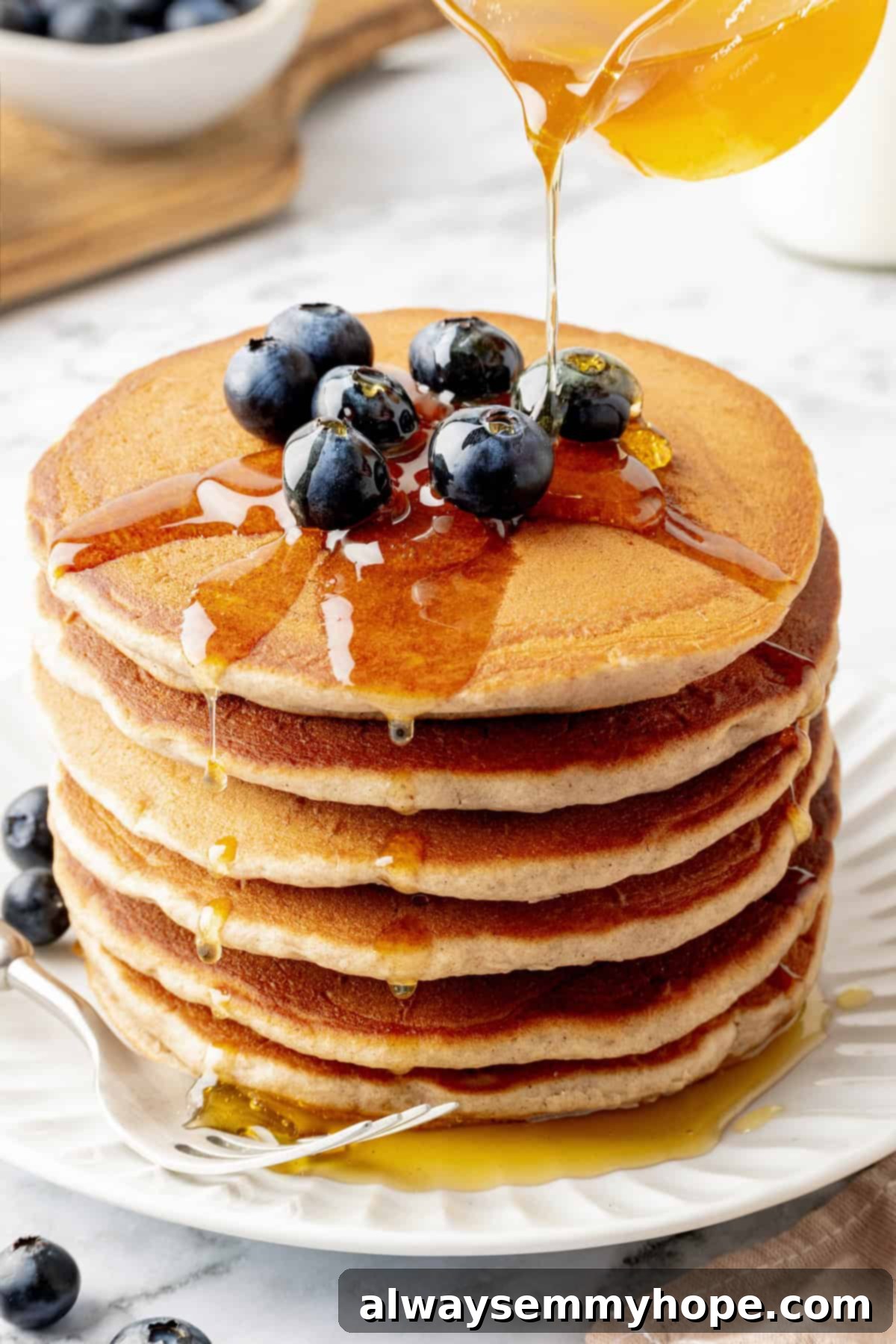 Hand pouring rich maple syrup over a tall stack of golden vegan buckwheat pancakes, creating a delicious cascade