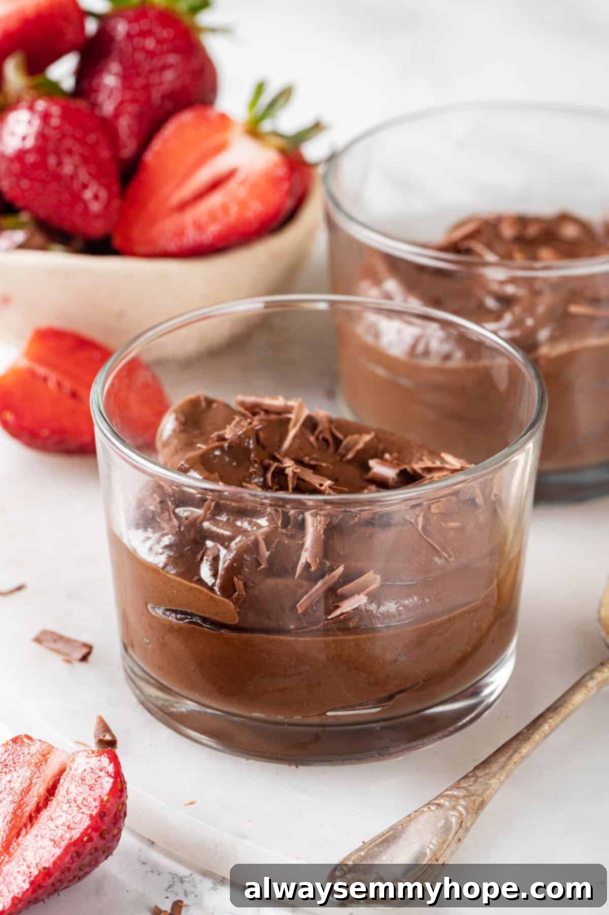 This easy avocado chocolate mousse recipe is rich, creamy, and oh-so-decadent—and you will never guess that the main ingredient is avocado! Two individual ramekins of chocolate avocado mousse, artfully garnished with chocolate shavings on top, ready for a luxurious dessert.