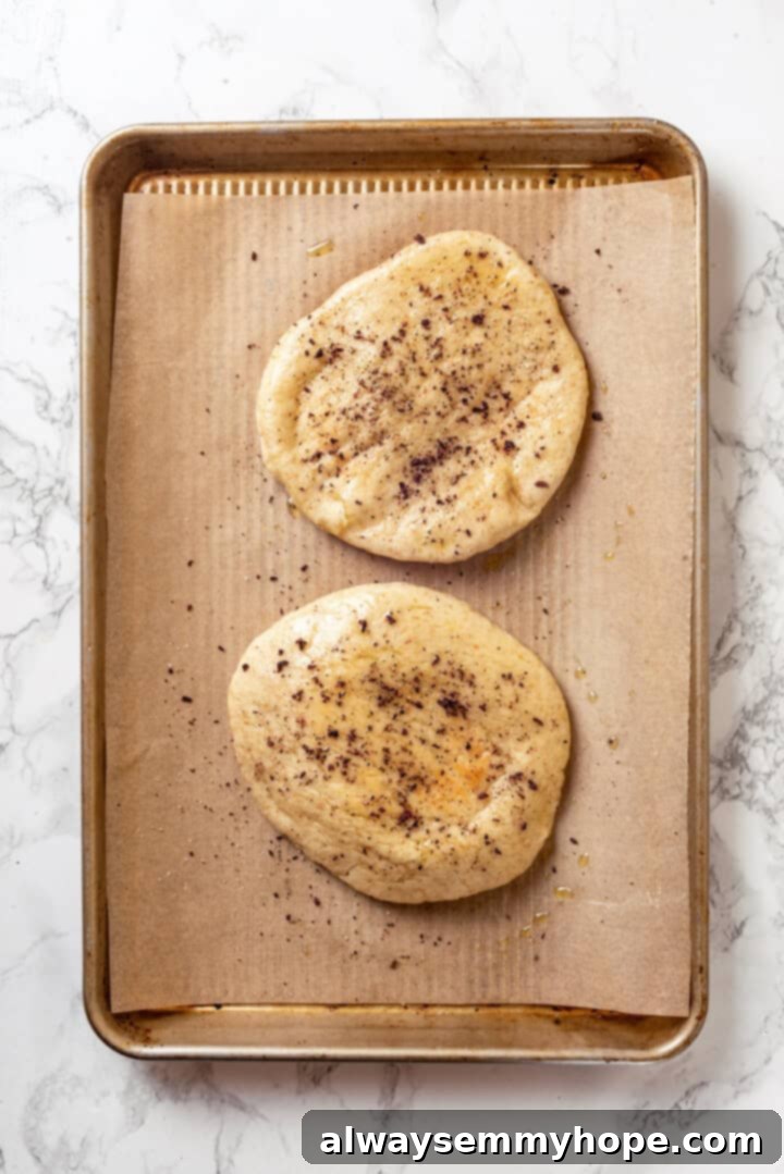 Pita bread brushed with olive oil and seasoned with sumac, salt, and pepper on a baking sheet. Seasoned and oiled pitas on baking sheet, ready for baking