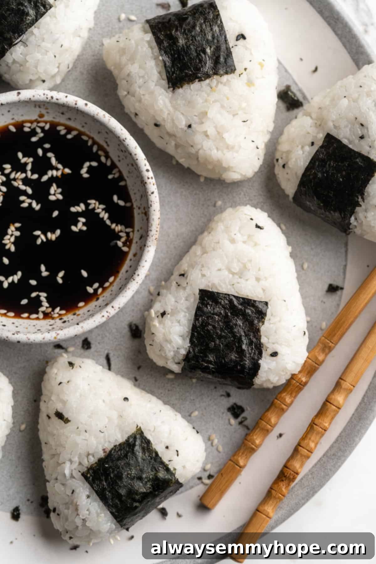 A beautifully arranged plate featuring several triangular vegan onigiri with chickpea tuna filling, garnished with nori strips, alongside a small bowl of savory dipping sauce. Perfect for a healthy snack or light lunch.