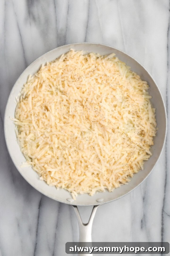 Overhead view of shredded potatoes seasoned with onion powder, salt, and pepper, forming an even layer in a hot skillet.