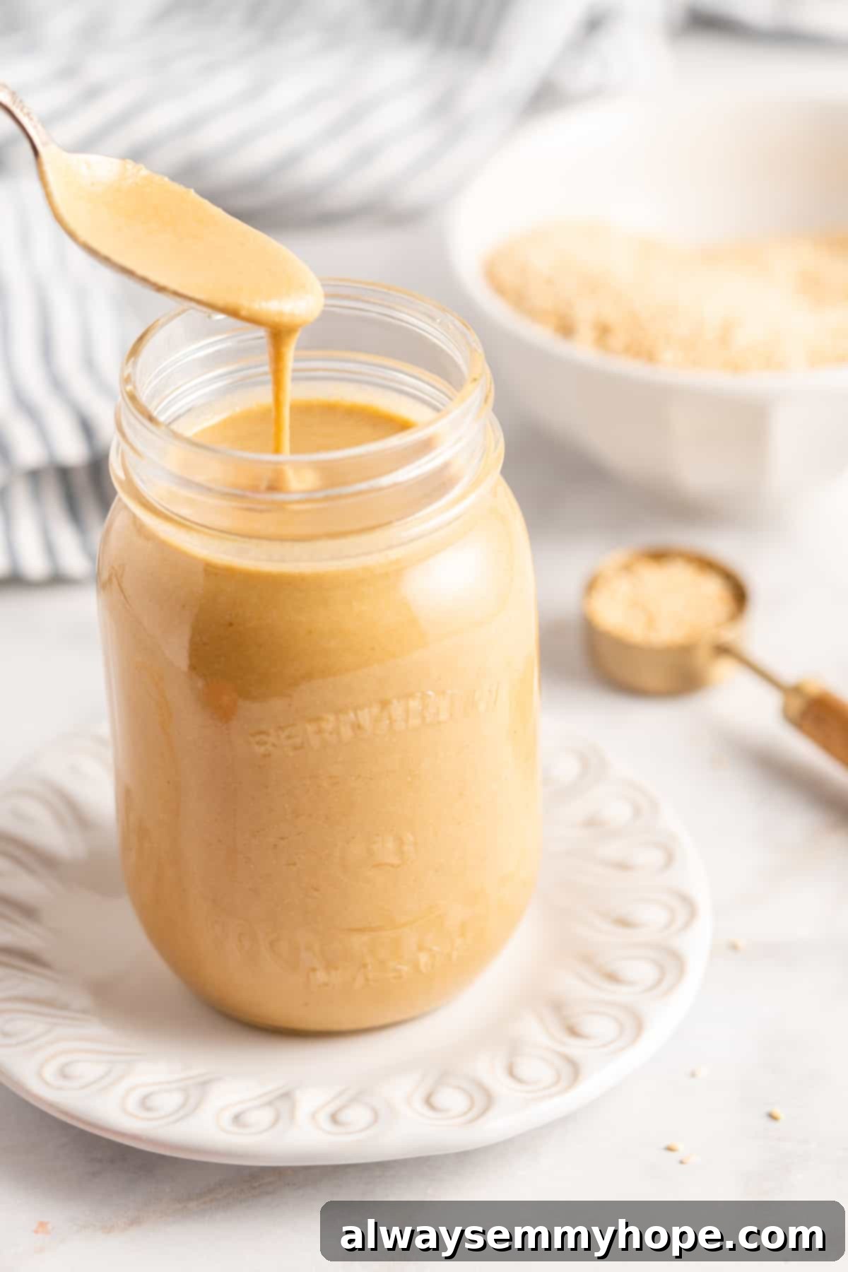 You’re two ingredients away from homemade tahini! Make it yourself and you’ll save so much money. Use in hummus, salad dressings, and more! A spoon overflowing with creamy homemade tahini is gracefully held over a glass jar, ready to be added, highlighting its rich texture and inviting appearance. The focus is on the golden-brown tahini, suggesting its freshness and quality.