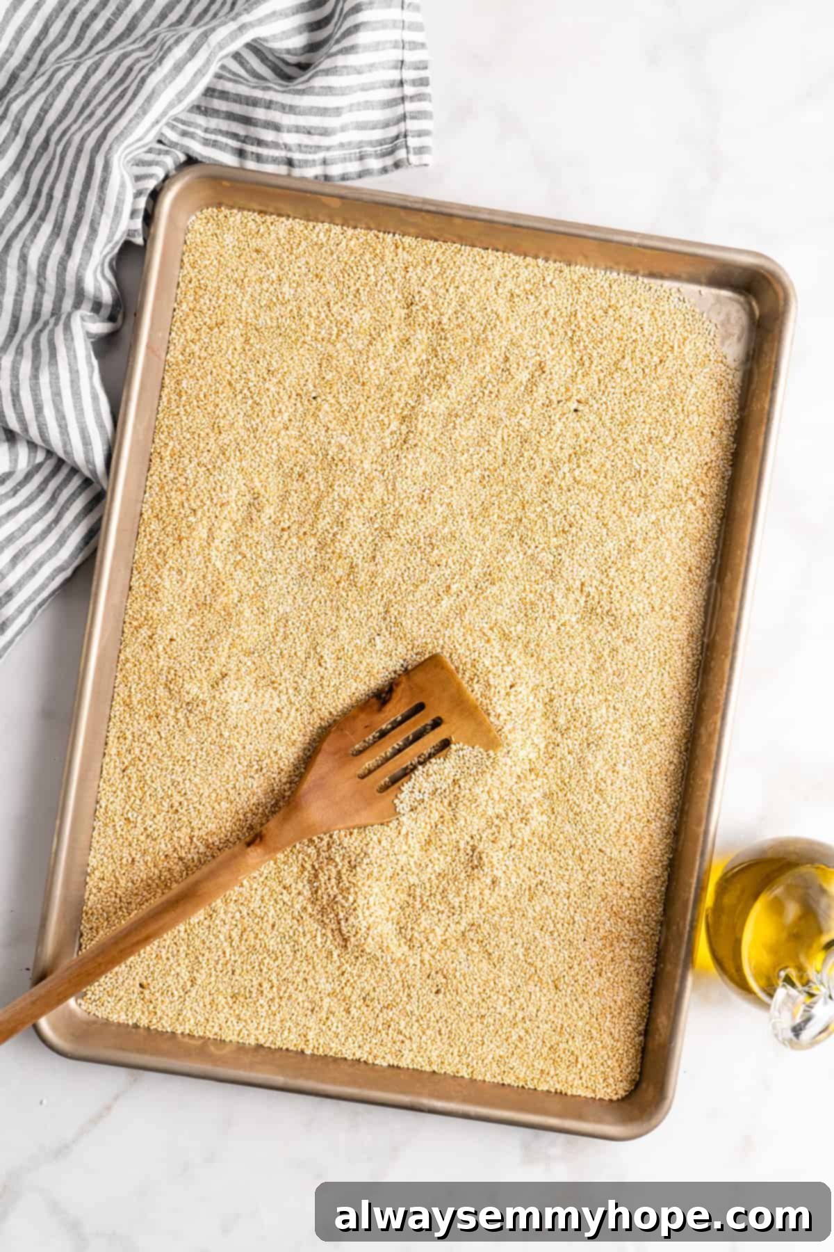 You’re two ingredients away from homemade tahini! Make it yourself and you’ll save so much money. Use in hummus, salad dressings, and more! An overhead view shows golden-brown toasted sesame seeds spread evenly on a rimmed baking sheet, with a wooden spatula resting alongside, suggesting readiness for the next step in tahini preparation.