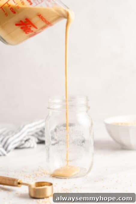 You’re two ingredients away from homemade tahini! Make it yourself and you’ll save so much money. Use in hummus, salad dressings, and more! A measuring cup full of creamy tahini is being poured into a clean glass jar, showcasing the smooth texture of the homemade sesame paste ready for storage or use.