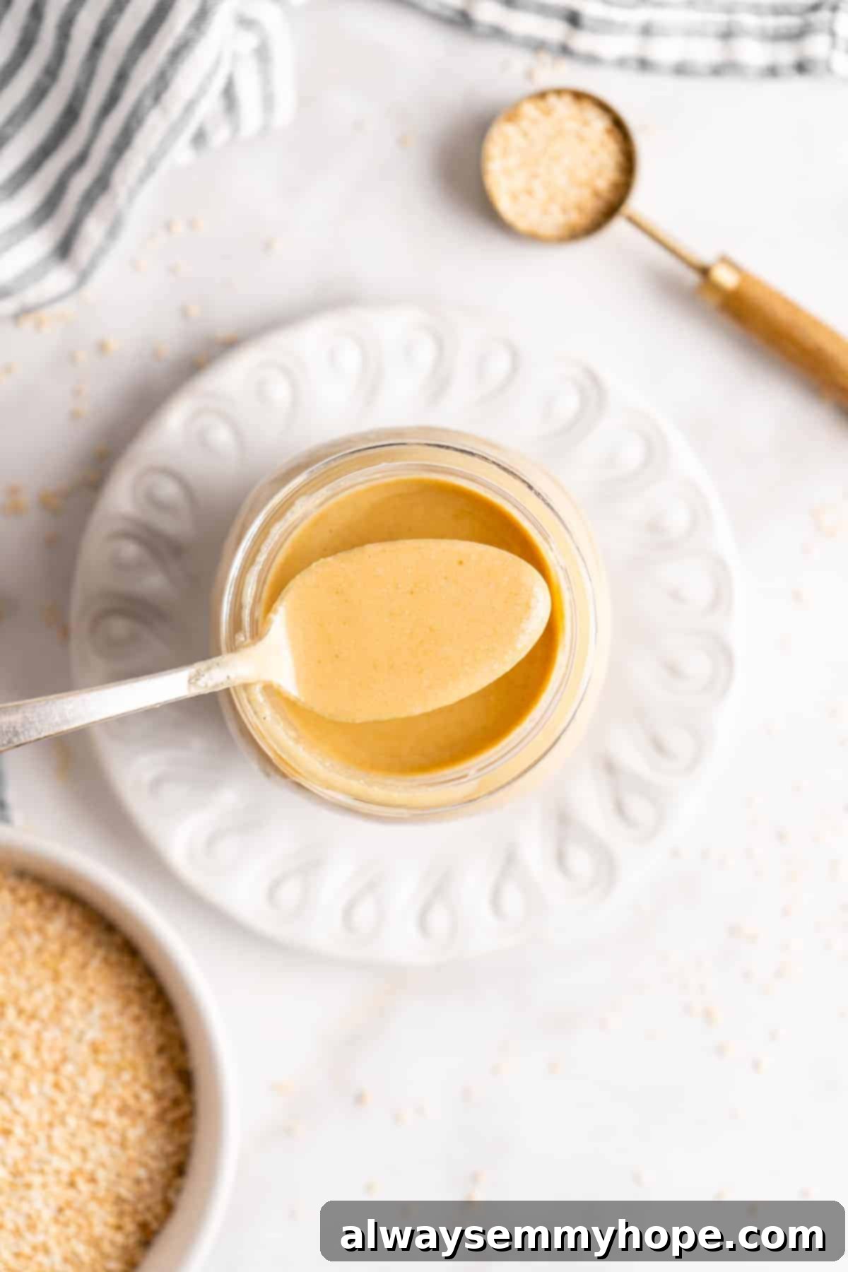 You’re two ingredients away from homemade tahini! Make it yourself and you’ll save so much money. Use in hummus, salad dressings, and more! An overhead view of a spoonful of creamy tahini being carefully held over a clear glass jar filled with more tahini, emphasizing its rich, flowing texture and readiness for use.