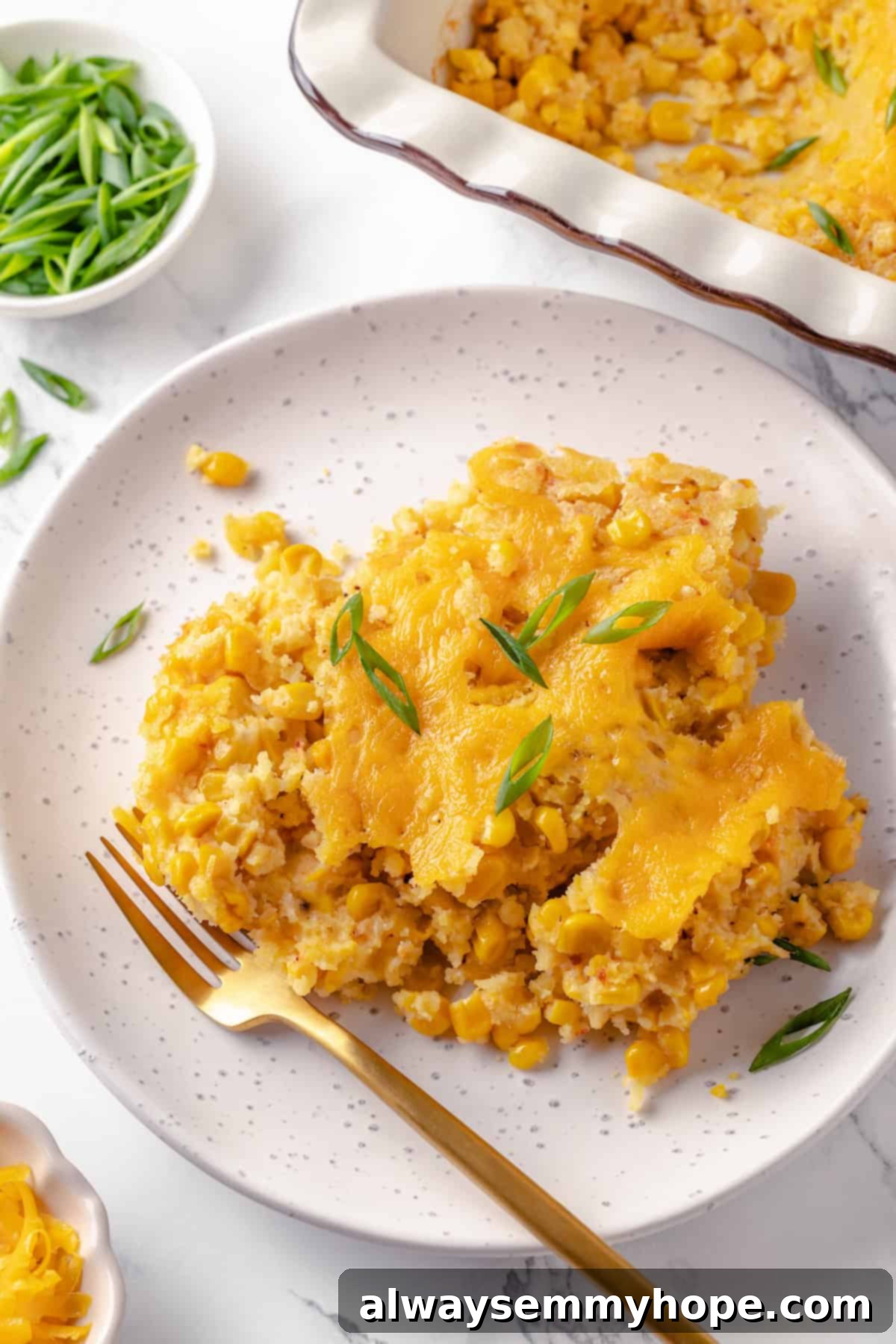 This easy vegan corn casserole is creamy, cheesy and sweet, with all the flavour you love of the holiday classic. Always a crowd-pleaser! A serving of creamy vegan corn casserole with melted cheese, garnished with fresh green onions, on a white plate with a fork.