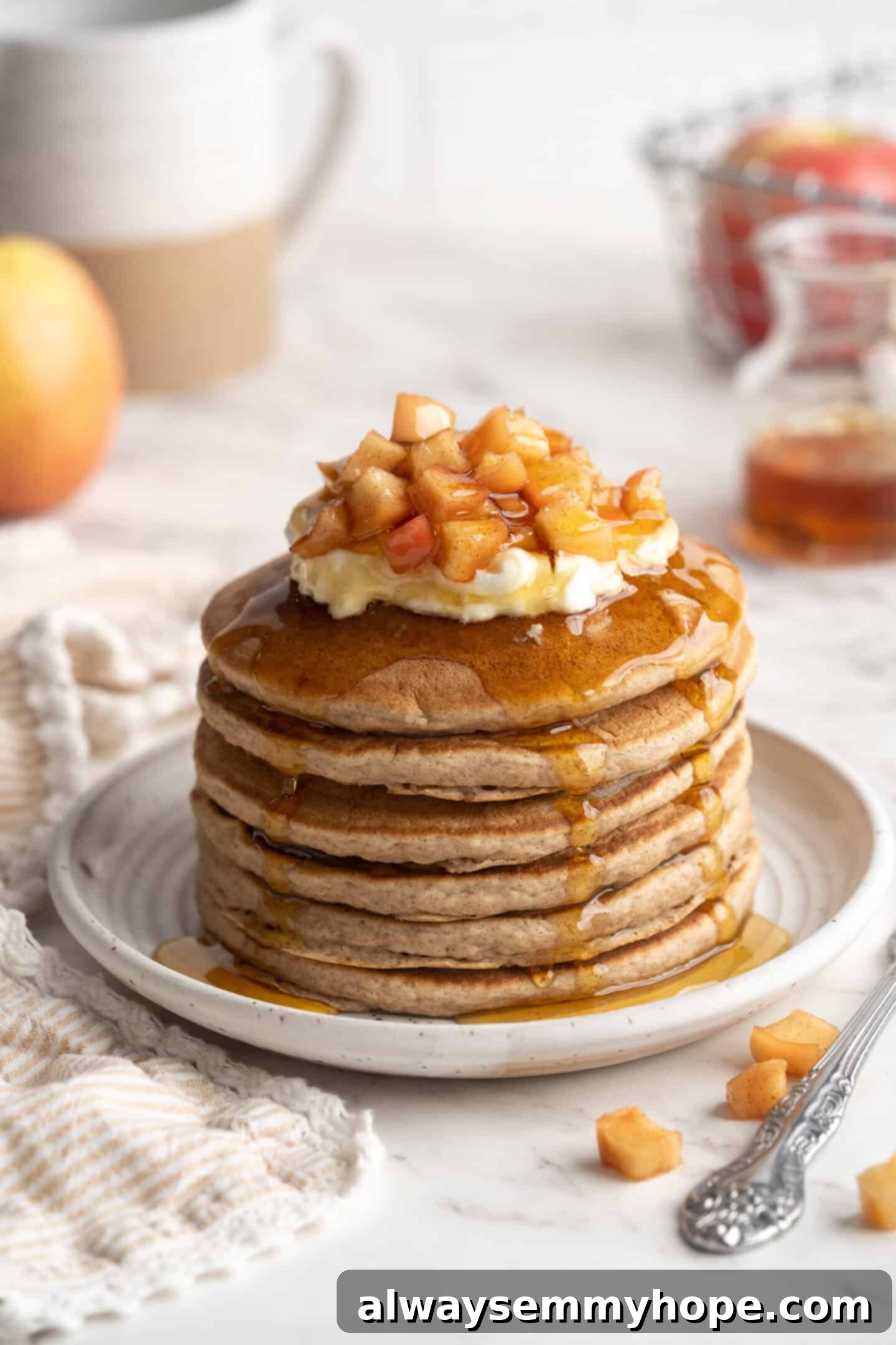 Stack of golden-brown apple pie pancakes on a white plate, adorned with a dollop of vegan yogurt, a drizzle of maple syrup, and fresh apple slices. The warm, inviting presentation hints at a delicious and wholesome breakfast.
