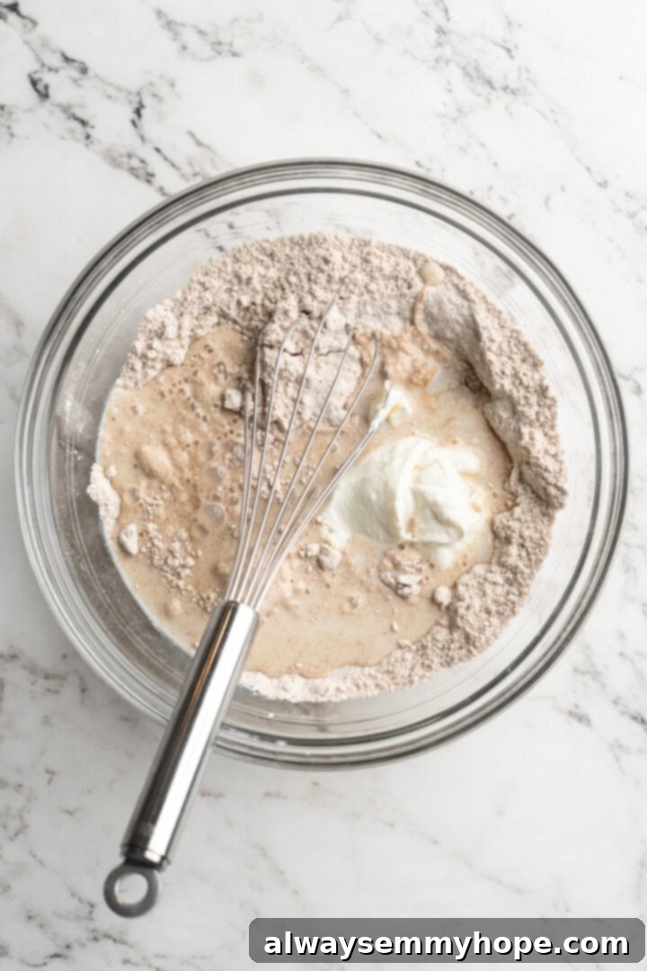 A whisk vigorously mixing light-colored pancake batter in a large mixing bowl, showing the consistency of the liquid ingredients combining.