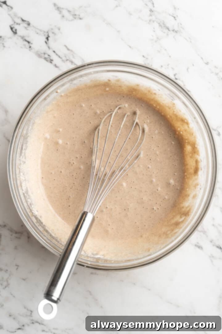 Close-up of a mixing bowl filled with smooth, pale yellow pancake batter, with a whisk resting inside, ready for the next step in preparing vegan apple pie pancakes.