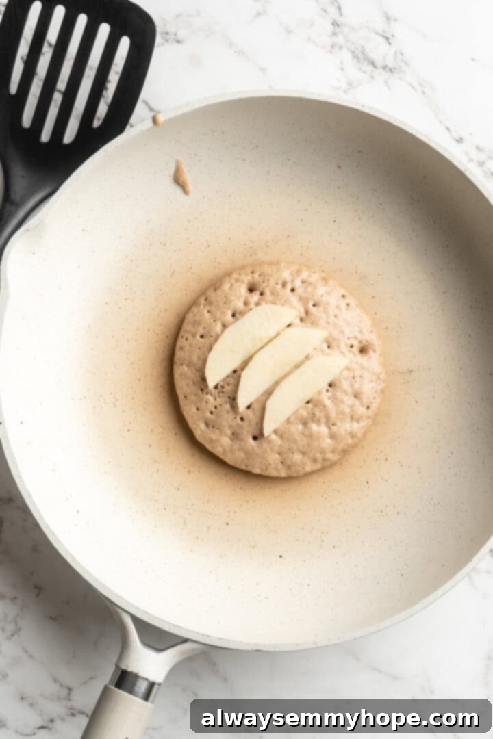 Several thin slices of fresh apple carefully placed onto the surface of a cooking pancake on a hot griddle, illustrating the step of adding fruit to the vegan apple pie pancake.