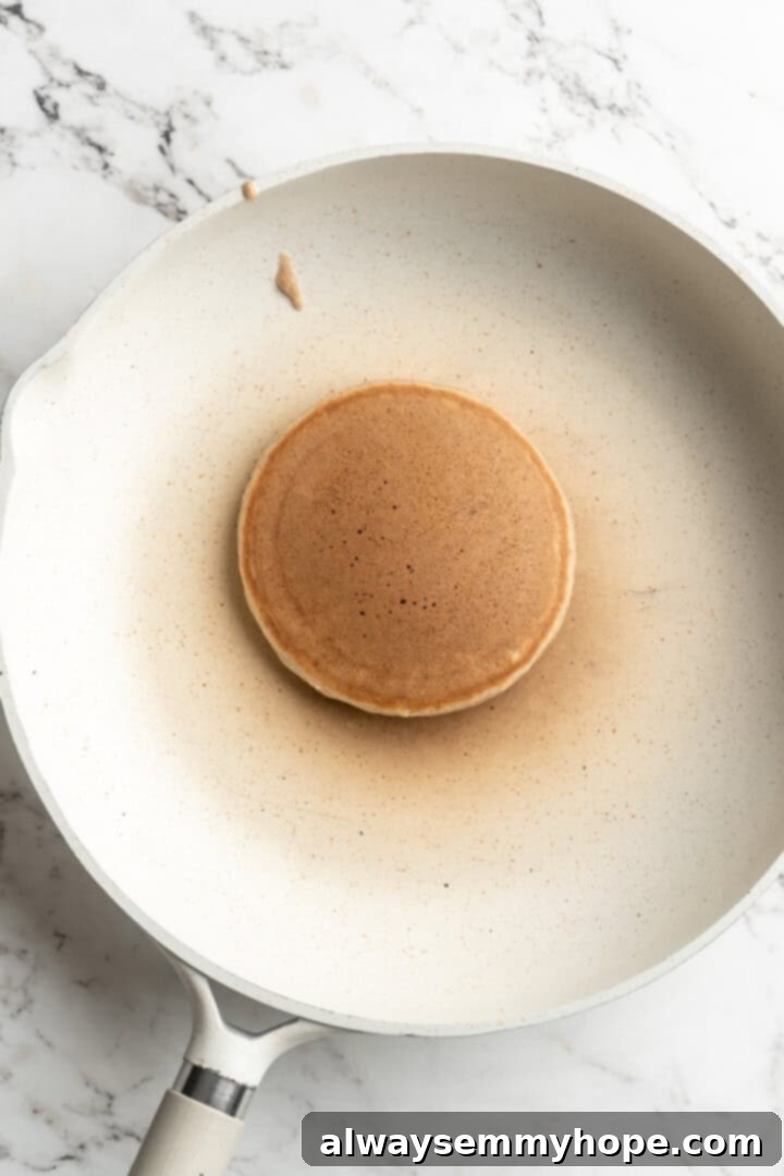 An overhead shot of a single apple pie pancake cooking on a hot griddle, with visible bubbles forming on the surface and delicate apple slices nestled within the batter, signaling it's nearly time to flip.