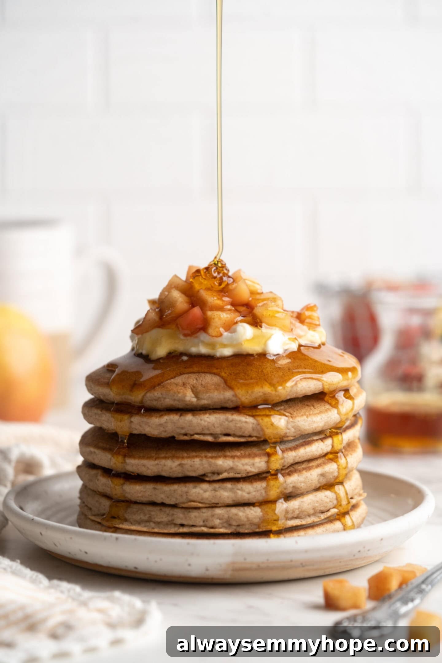 A close-up shot of rich maple syrup being drizzled over a towering stack of golden vegan apple pie pancakes, highlighting the sweet, glossy texture of the syrup and the soft texture of the pancakes beneath.